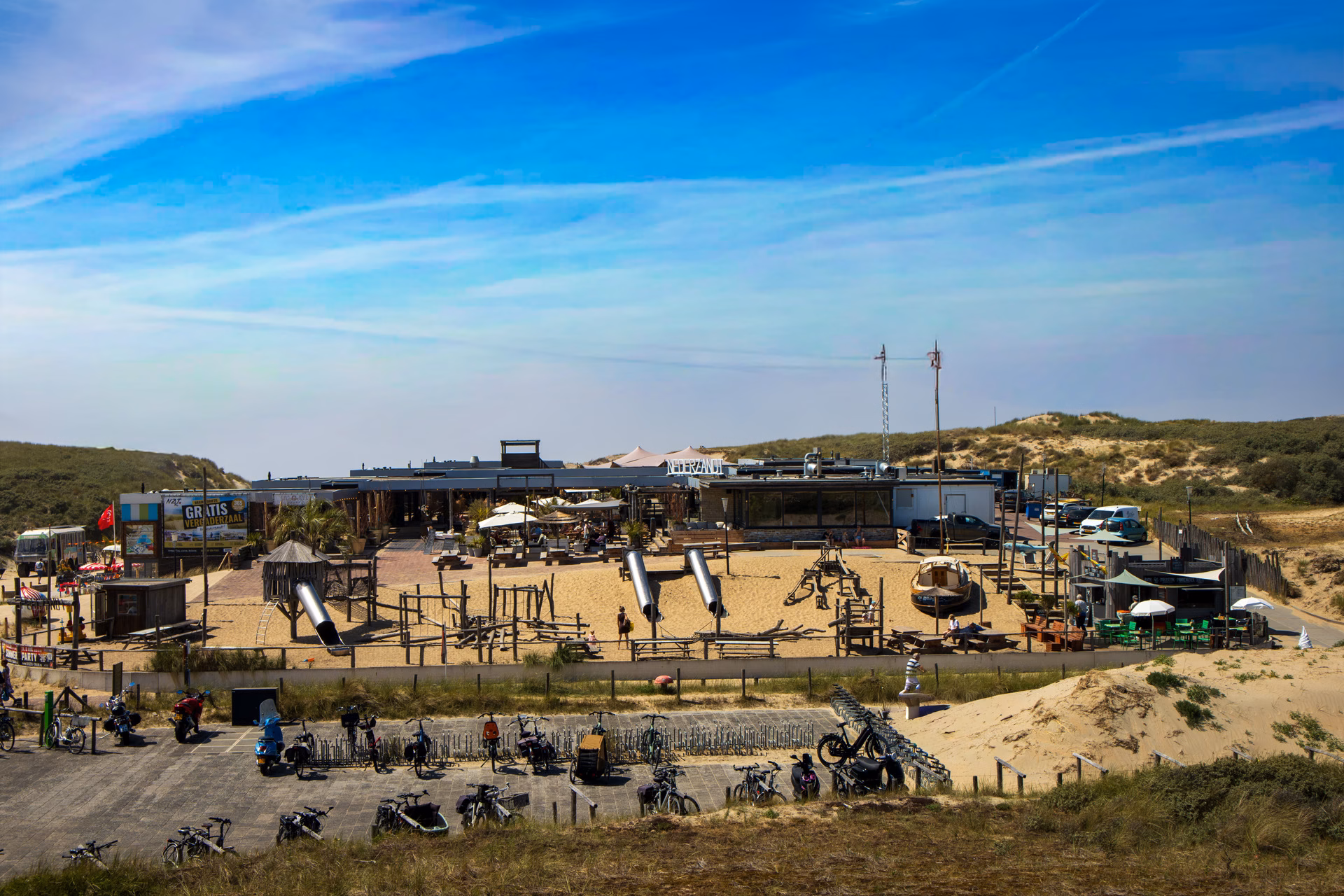 Groot familievriendelijk strandpaviljoen met speelstrand, glijbanen, terrassen en fietsenstalling tussen de duinen onder een helderblauwe lucht
