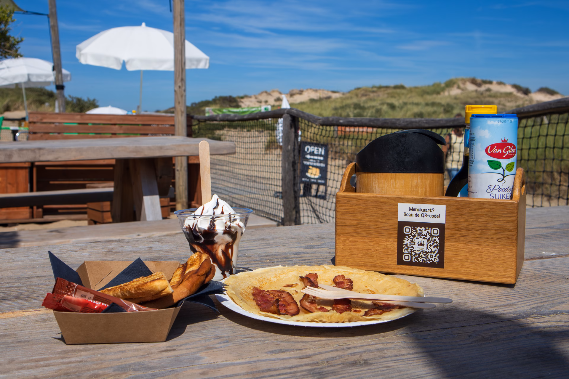 Pannenkoek met spek, tosti en softijs op houten picknicktafel met uitzicht op duinen en strandterras; menukaart via QR-code op tafel.